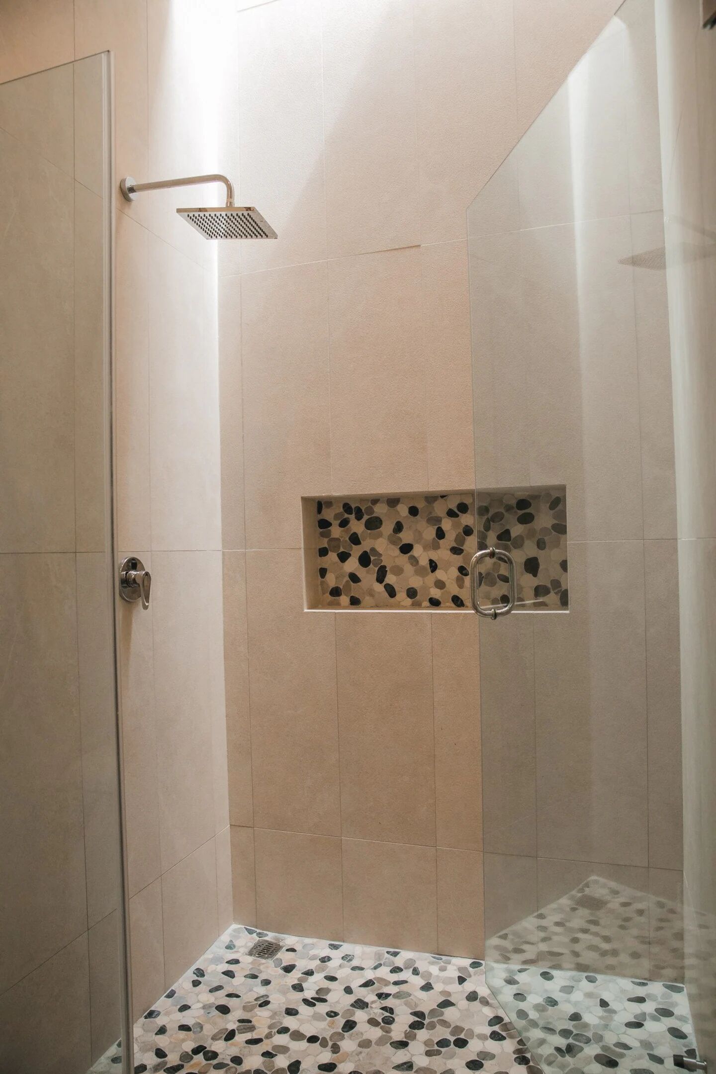 A shower with a glass door, a pebble-patterned floor, and a wall-mounted shelf.