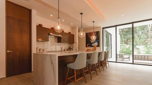 A modern kitchen with a marble island, bar stools, pendant lights, and a large window with a view of greenery.