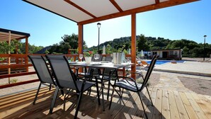 Un espace extérieur aménagé pour les repas, avec une table dressée pour deux personnes, entourée de chaises, sous une pergola en bois. On aperçoit une piscine et des chaises longues à l’arrière-plan.