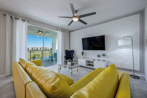 Uma sala de estar moderna com um sofá amarelo, uma mesa de centro de vidro, um ventilador de teto, uma televisão de tela plana e vista para o exterior.