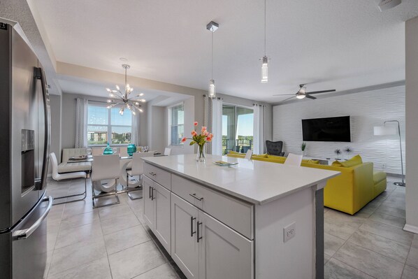 A modern kitchen with a central island, dining area with a sofa, and a living room with a TV and yellow sofa.