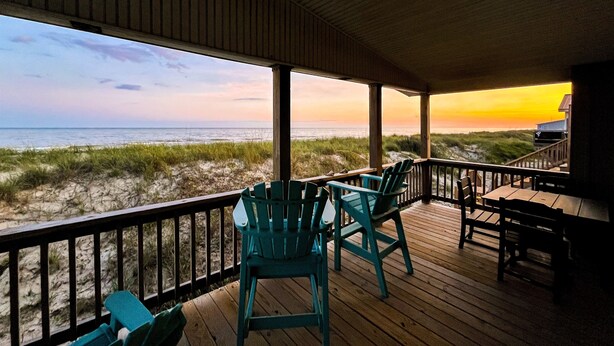 A covered deck with chairs overlooking a beach at sunset. - Salt Life - charming beachfront cottage! Steps to the beach! (Oak Island)