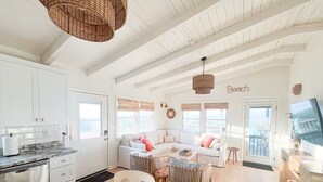 A bright, white interior with a kitchen, dining area, and living room. There are wicker baskets, a white sofa, and a television. - Salt Life - charming beachfront cottage! Steps to the beach! (Oak Island)