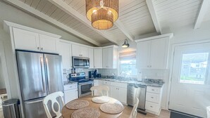 A modern kitchen with white cabinets, stainless steel appliances, and a dining area with a wooden table and white chairs. - Salt Life - charming beachfront cottage! Steps to the beach! (Oak Island)