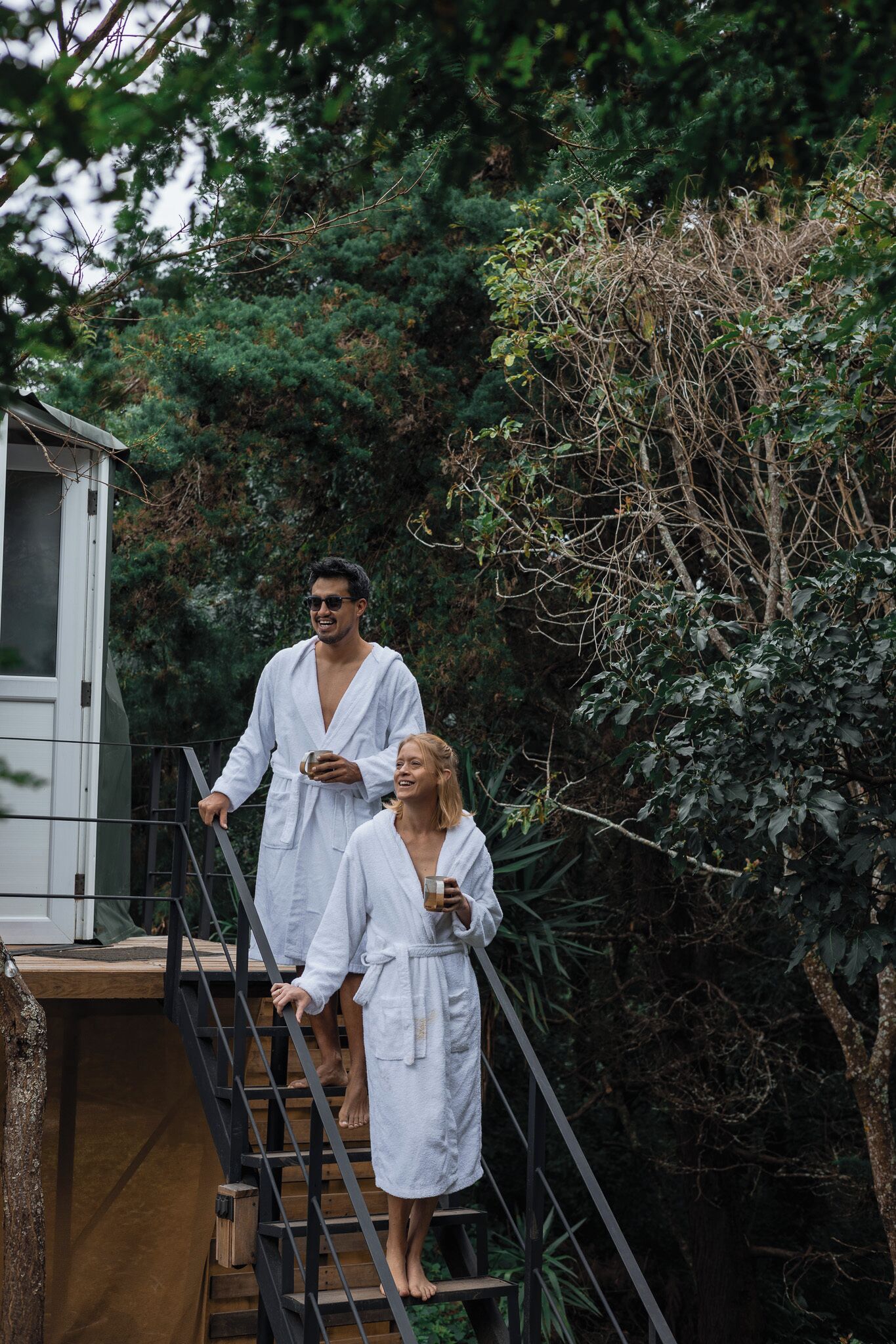 Two people in white bathrobes descending a wooden staircase with metal railings, surrounded by dense greenery.
