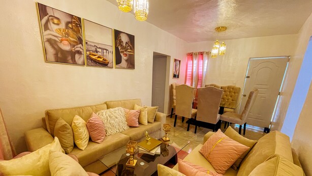 Een woonkamer met een beige bank, bijpassende fauteuils en een salontafel. Er hangen afbeeldingen in lijsten aan de muur en een kroonluchter erboven.
