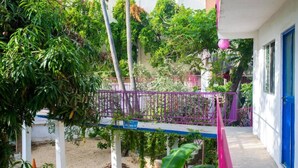 A covered outdoor area with a purple metal railing, surrounded by greenery and a white building with a blue door.