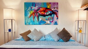 A bed with a patterned blue cover, four pillows, a purple throw, and a colorful abstract painting above.