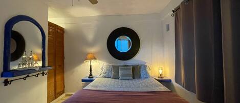 A bedroom with a bed, a ceiling fan, a round mirror, a nightstand with a lamp, and a window with curtains.