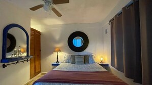 A bedroom with a bed, a ceiling fan, a round mirror, and a nightstand with a lamp.