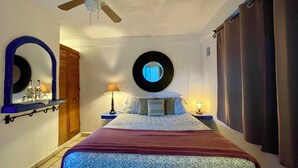 A bedroom with a bed, a ceiling fan, a round mirror, a nightstand with a lamp, and a window with curtains.