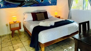 A bedroom with a bed, two bedside lamps, a ceiling fan, a window with curtains, and a painting of dolphins.