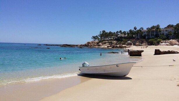 A beach with a boat on the sand, clear blue water, and a rocky shoreline with buildings in the background.