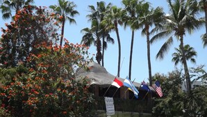A building with a thatched roof, surrounded by palm trees and red flowers. Flags are displayed on the building.