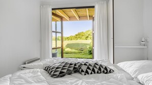 Un dormitorio con un ventanal grande, una cama con ropa blanca y almohadas estampadas, y una mesita de noche blanca con una lámpara.