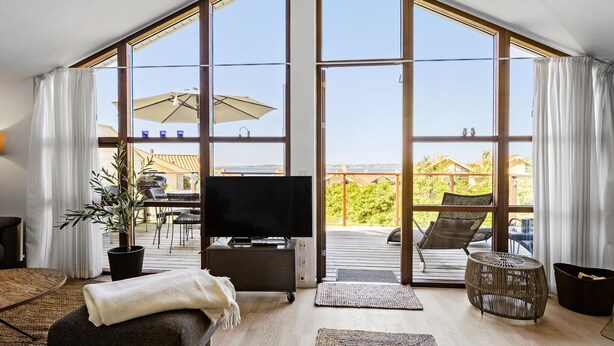 Un salón amplio con un televisor grande, asientos cómodos, un balcón con mobiliario exterior y vistas al mar.