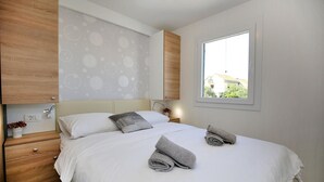Ein ordentlich bezogenes Bett mit weißen Leinen und grauen Handtüchern, ein Nachttisch mit einer Pflanze und ein Fenster mit Blick auf Gebäude.