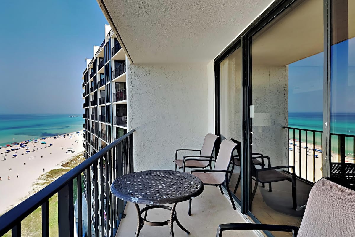 Een balkon met een tafel en stoelen, met uitzicht op een strand met parasols en mensen.