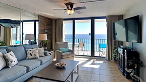 A living room with a sofa, coffee table, and a TV. There is a balcony with a view of the ocean. - Edgewater Tower III by Resort Collection (Panama City Beach)