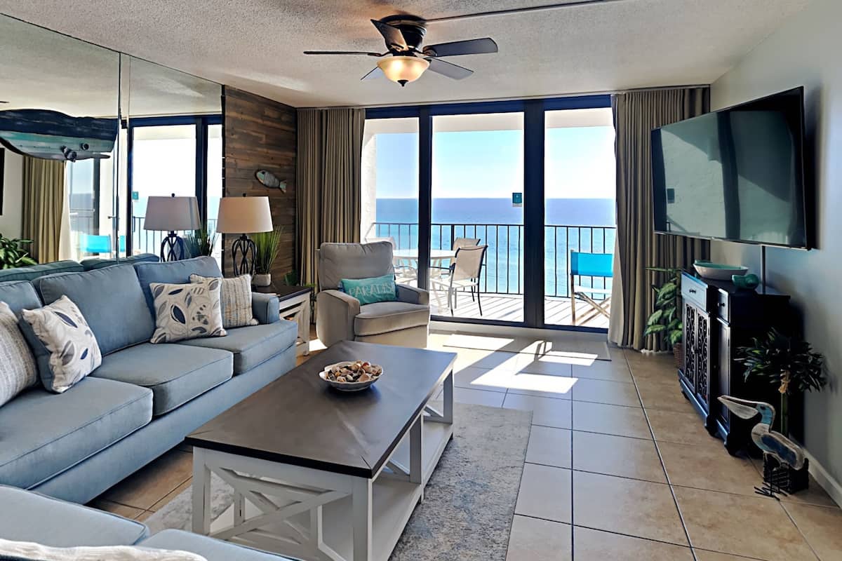 Een woonkamer met een bank, salontafel en een televisie. Er is een balkon met uitzicht op de oceaan.
