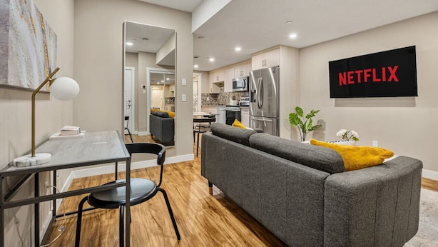A modern living room with a grey sofa, a dining table with chairs, a TV mounted on the wall, and a kitchen area in the background. - Cozy & Comfortable 2 Bedroom Suite, Mins to Airport, NW Calgary. (Calgary)