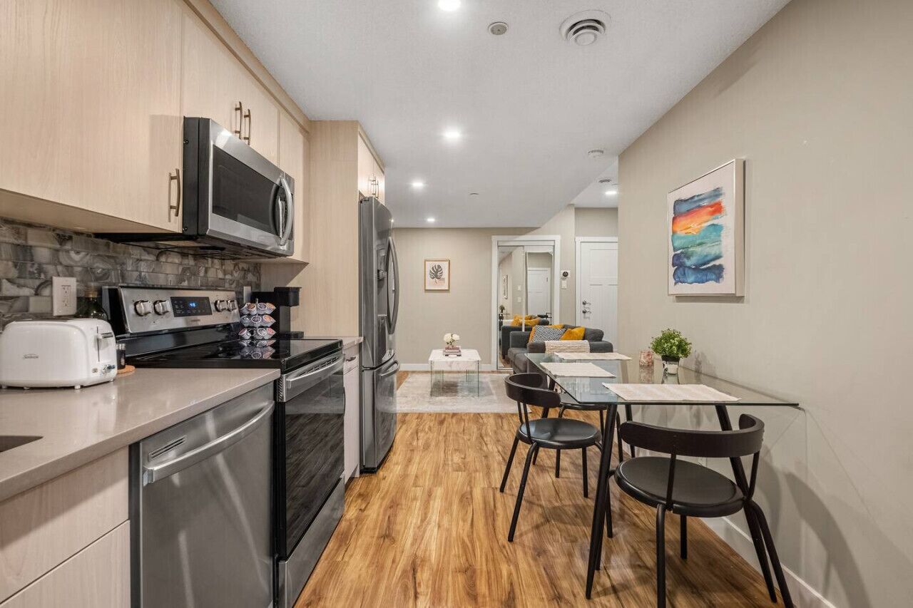 A modern kitchen with stainless steel appliances, a dining area with a glass table, and a living space visible through an open door.