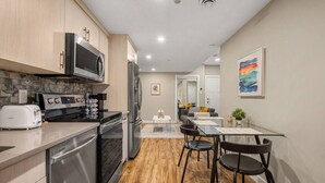 A modern kitchen with stainless steel appliances, a dining area with a glass table, and a living space visible through an open door. - Cozy & Comfortable 2 Bedroom Suite, Mins to Airport, NW Calgary. (Calgary)
