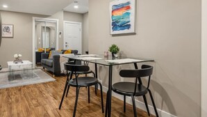 A modern living room with a glass dining table, black chairs, a sofa with cushions, a coffee table, and a painting on the wall. - Cozy & Comfortable 2 Bedroom Suite, Mins to Airport, NW Calgary. (Calgary)