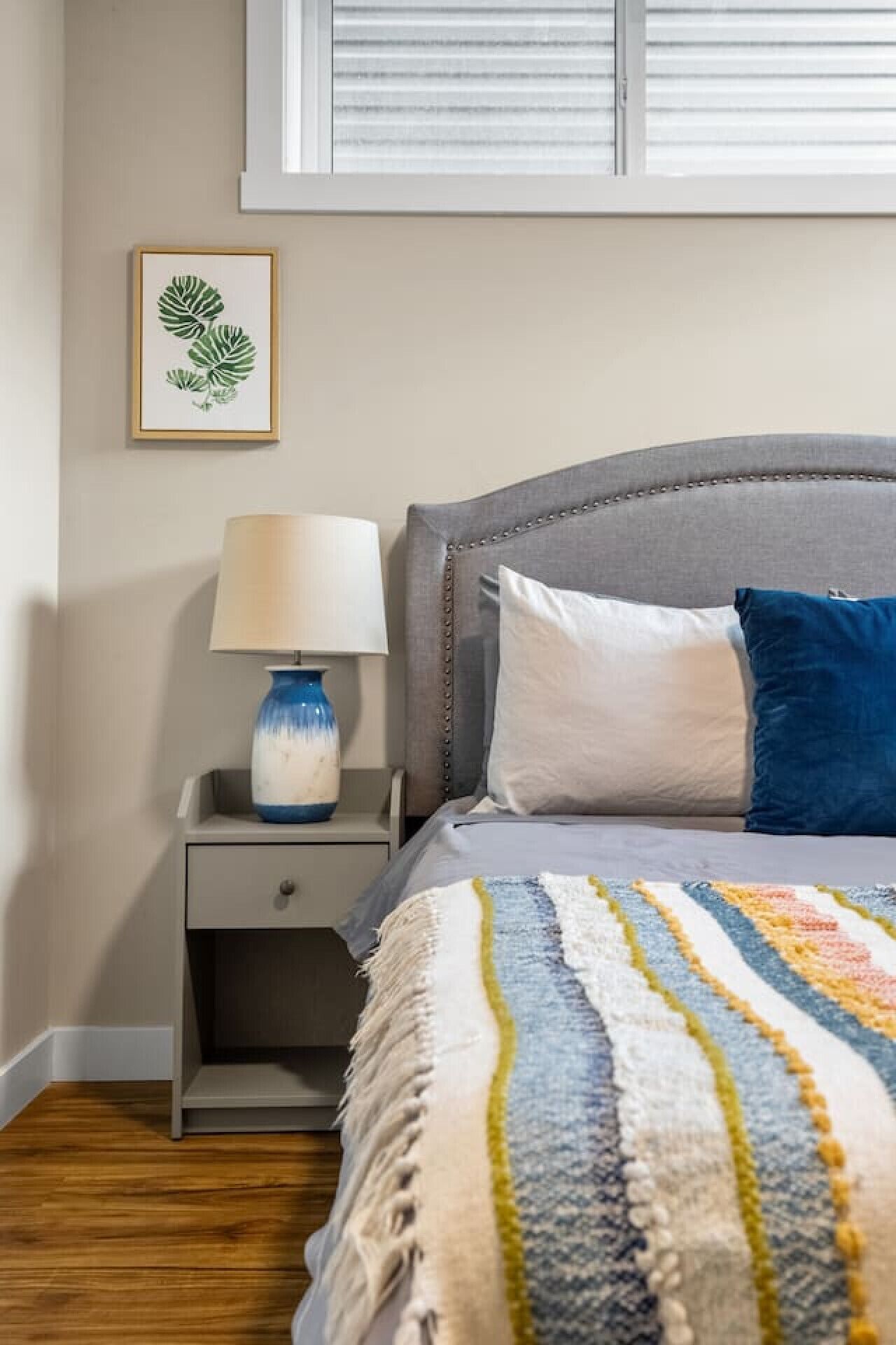 Een slaapkamer met een bed, een nachtkastje, een lamp en een plant in een lijst.