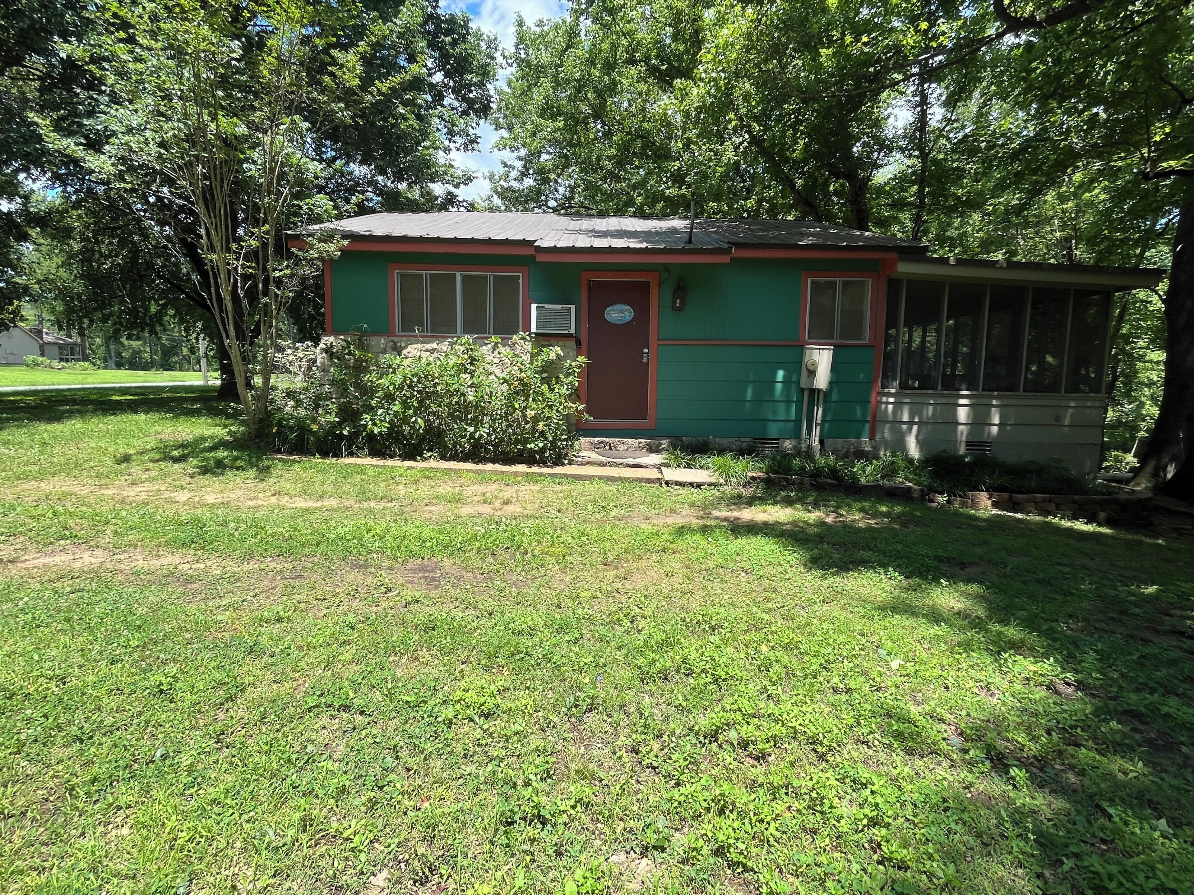 A small, single-story building with a green exterior and a red door, surrounded by a grassy yard and trees.