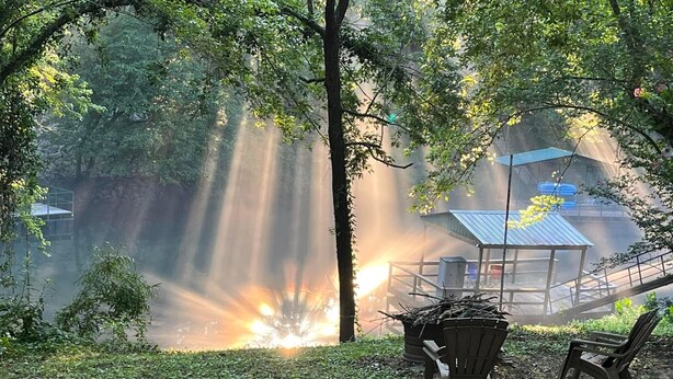A serene outdoor area with a bench and a chair, surrounded by lush greenery and a view of a distant fire. - Cozy family fish camp on the Little Red River, a premier trout fishing stream! (Pangburn)