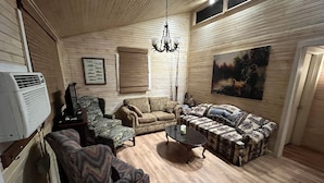 A living room with a wooden floor, a sofa set, a patterned armchair, a small coffee table, a wall-mounted air conditioner, and a painting. - Cozy family fish camp on the Little Red River, a premier trout fishing stream! (Pangburn)