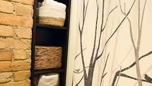 A bathroom with a shower curtain featuring a tree design, a brick wall, and shelves with baskets and towels.