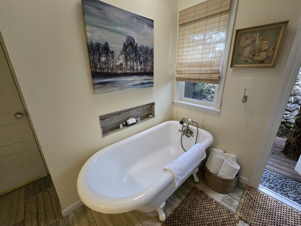 A bathroom with a freestanding bathtub, a window with blinds, a painting above the bathtub, and a small shelf with toiletries. - Cozy Volcano Cottage (Volcano)