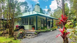 A small, single-story building with a green roof and a covered porch, surrounded by lush vegetation and a gravel driveway. - Cozy Volcano Cottage  (Volcano)