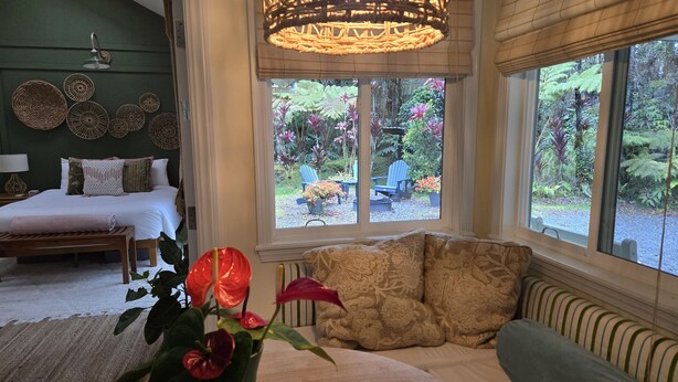 A bedroom with a bed, bedside table, lamp, and a view of a garden through large windows. - Cozy Volcano Cottage  (Volcano)