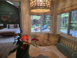 A bedroom with a bed, bedside table, lamp, and a view of a garden through large windows. - Cozy Volcano Cottage (Volcano)