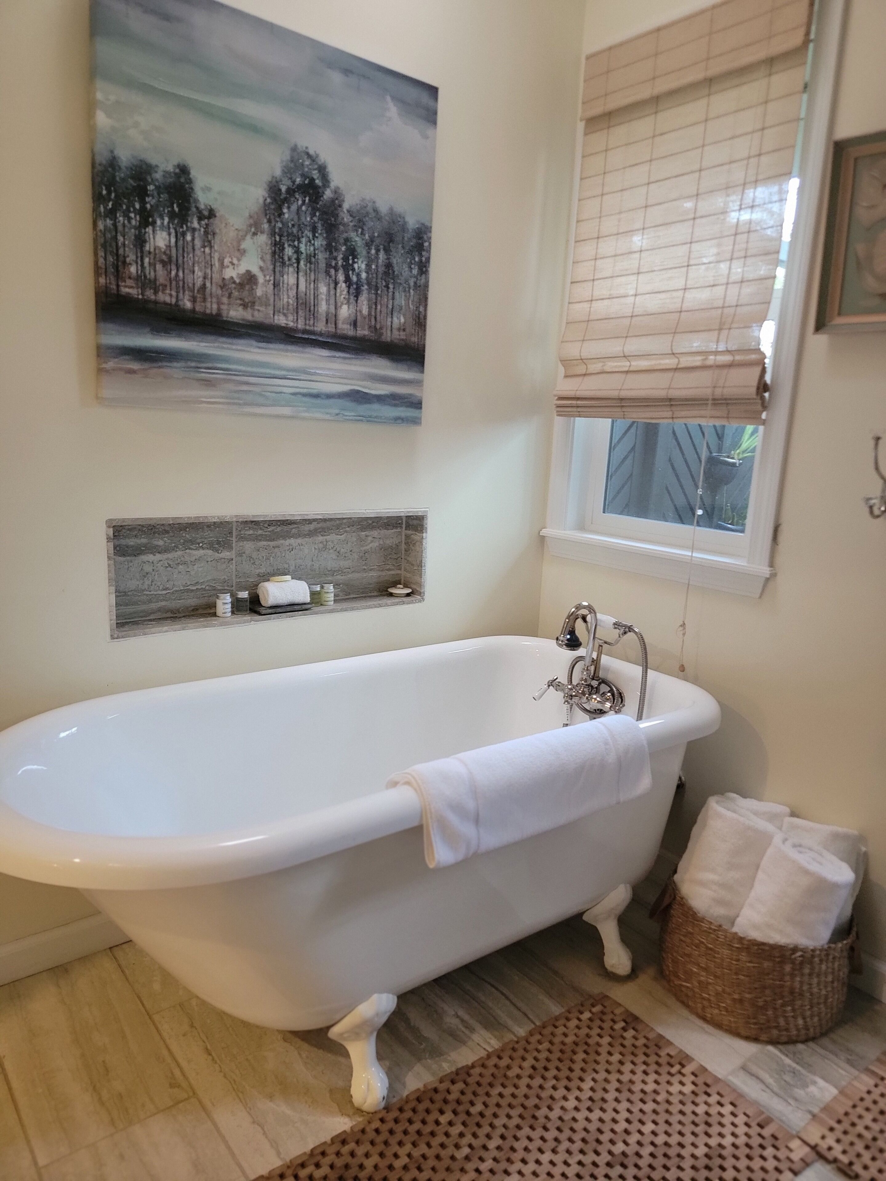 A bathroom with a freestanding bathtub, a window with blinds, a painting on the wall, and a shelf with toiletries.