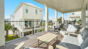 Une terrasse extérieure couverte, dotée d’une table en bois et de sièges blancs rembourrés, qui offre une vue sur un quartier résidentiel avec des maisons blanches et un ciel bleu dégagé.