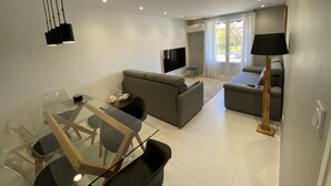 A modern living room with a grey sofa, a glass dining table, and a TV mounted on the wall. - Le Coup de Cœur Dargelès (Argelès-sur-Mer)