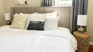 A neatly made bed with white bedding and patterned pillows, a tufted headboard, and a woven side table with a lamp.