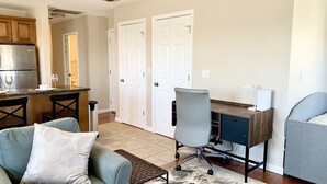 A modern living room with a gray sofa, a coffee table, and a desk with a chair. There is a kitchen area with a refrigerator and a dining table with chairs. The room has white doors and a tiled floor.