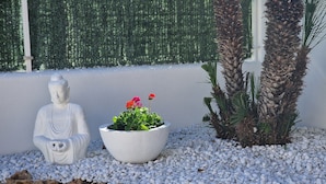 A Buddha statue, a potted plant with flowers, and a palm tree in a landscaped area with white pebbles and rocks. - "Gecko"@MARISOL: 250m to the beach, private pool, A/C, WiFi, dog beach (Riumar)
