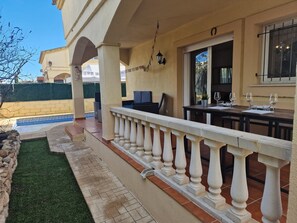 Eine überdachte Terrasse im Freien mit Pool, einer Essgegend mit Tisch und Stühlen sowie einem kleinen Garten mit Hecke und Steinmauer.