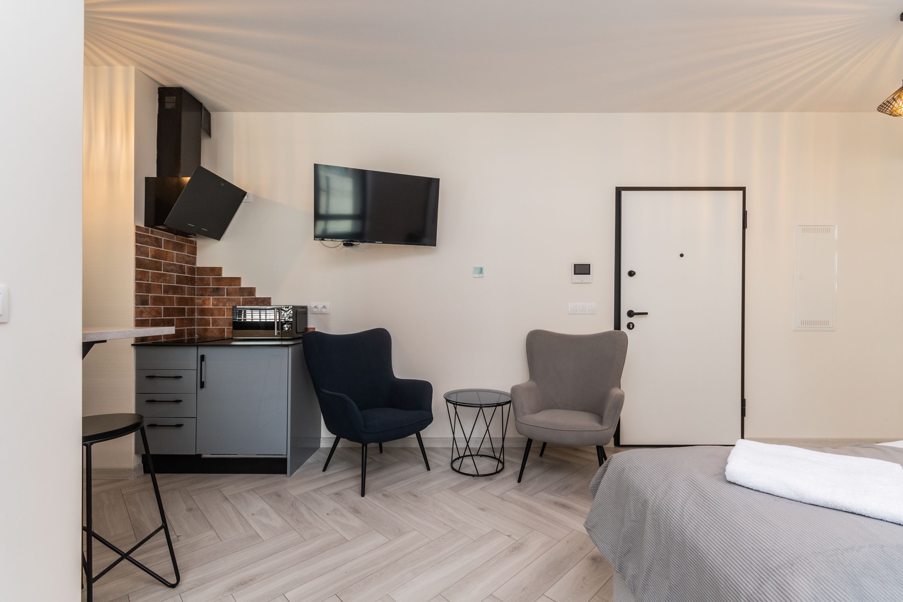 Habitación de hotel compacta con kitchenette, televisión de pantalla plana montada en la pared, cama con manta blanca y dos sillas.