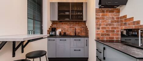 A compact kitchen with grey cabinets, a herringbone-patterned backsplash, and a black countertop.