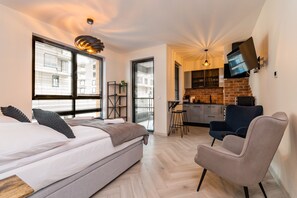 A modern bedroom with a bed, a chair, and a kitchen area visible through an open door. - Doki Studio Stocznia by Grand Apartments (Gdansk)