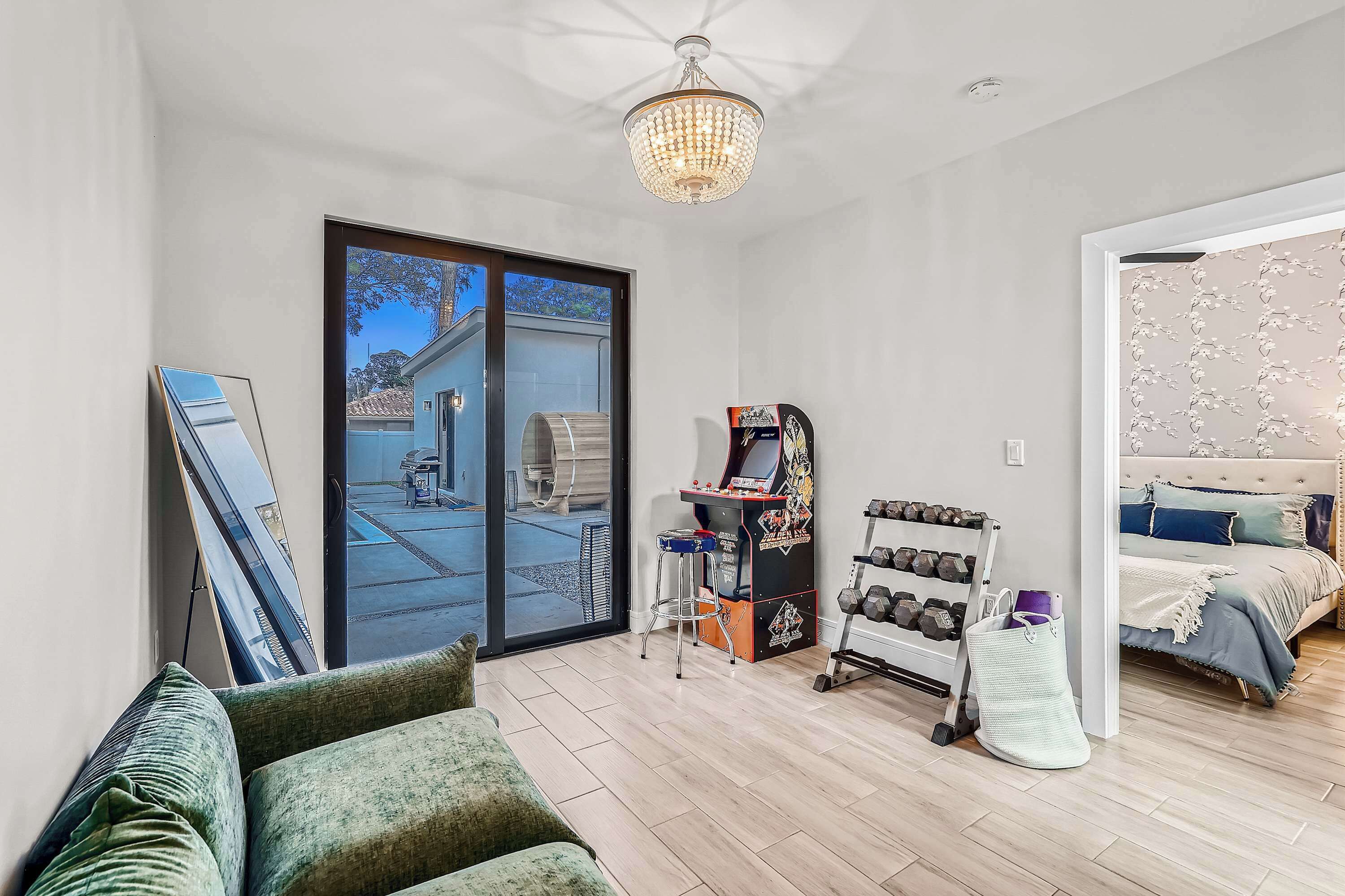 Ein modernes Zimmer mit einer großen Schiebeglasdoor zum Patio, einer Spielkonsole, einem Bett mit dekorativem Kopfteil und einem kleinen Fitnessbereich inklusive Kurzhanteln.