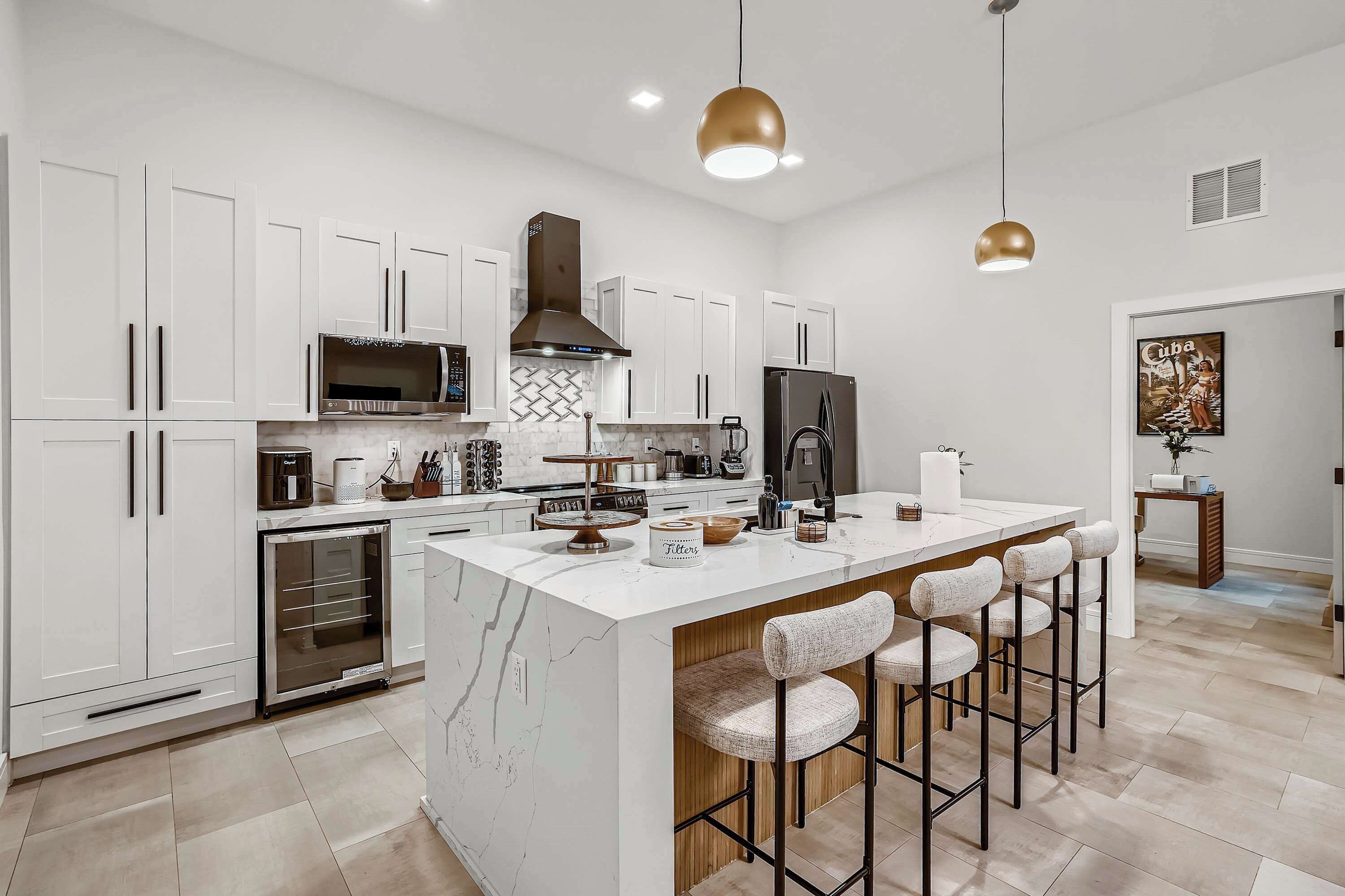 Uma cozinha moderna com armários brancos, uma ilha central com banquetas, eletrodomésticos de aço inoxidável e iluminação pendente.