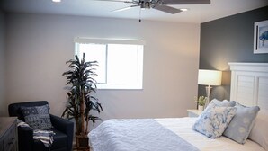Een slaapkamer met een bed, een stoel, een potplant, een nachtkastje en een plafondventilator.